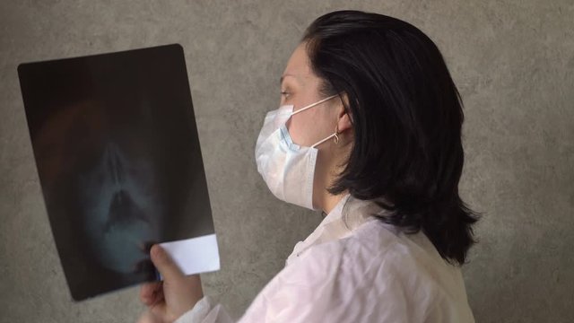 A young woman is wearing protective clothing. There is a medical mask on the face. Holds an x-ray of the head in his hands. He hits the picture with his hand. In room. Therapist. Consultant.