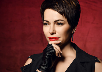 Portrait of brunette woman in leather gloves with cut fingers touching holding her chin over dark red background