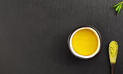 Cup with matcha green tea on a black stone background. Tea leaves and a wooden spoon with tea nearby. Top view, tea ceremony, minimalism, copy space for text.