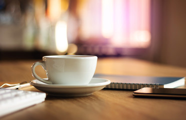 Closeup white cup of coffee with smartphone, notebook and computer keyboard on table in cafe. Vintage light, blurred and bokeh background