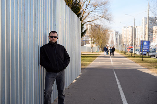 Street photo of a handsome man in a black jacket with glasses in the city at the crossroads on a sunny day near the wall