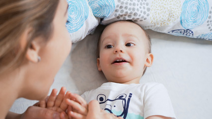 Beautiful young mother and toddler son enjoying each other in the morning. Smiling, laughing and playing in bed with child. Family happiness concept. Bright, sunny warm day.