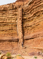a long vertical magmatic igneous dike cuts through red and orange horizontal layers of sedimentary...