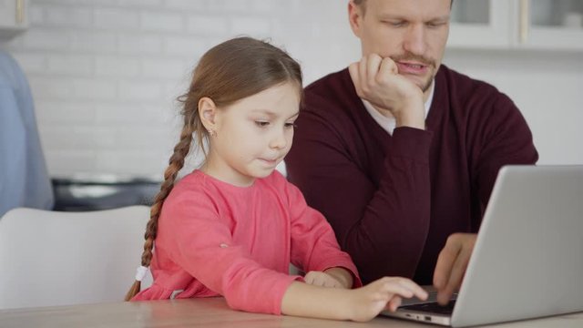 Tilt down medium shot of middle aged father teaching curious little daughter how to use laptop computer sitting at table at home