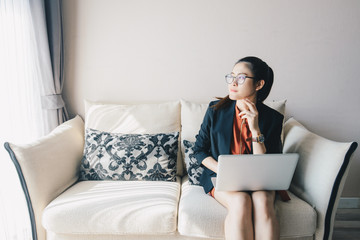 Portrait of young Asian businesswoman working online with her laptop and thinking something while sitting on a sofa in an office. Portrait of businesswoman while working concepts.