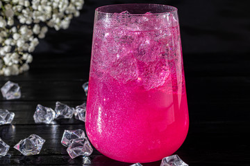 A sparkling brilliant raspberry drink in a glass stands on a wooden table with ice. In the...