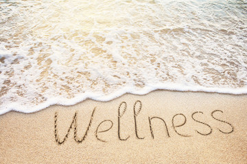 Wellness word written on sand. Beach, soft wave and warm light background.