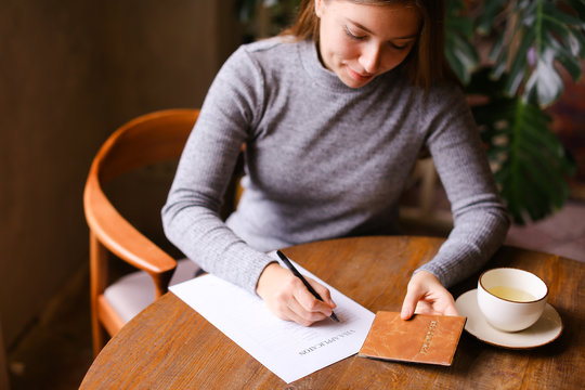 Young cauacsian girl filling visa application at cafe, passport on table.
