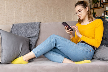 young girl in jeans an orange sweater and glasses sits on a sofa and looks at a smartphone