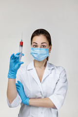 Young woman doctor in white uniform, mask and rubber gloves holding syringe on gray background.