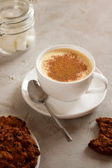 Cappuccino coffee in white cup with tea spoon and american cookies with chocolate drops and defocused sugar bowl on grey table. Cement texture. Vertical composition with copy space. Breakfast. Cafe.