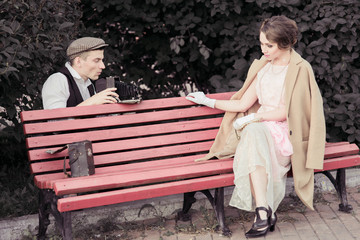 A young couple is photographed on a retro camera while walking in the Park. A woman sits on a bench , a man photographs a stylishly dressed woman in a retro style.Historical reconstruction