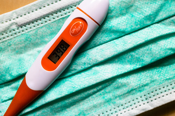Digital thermometer is laying medical face mask and displaying high temperature or fever reading. Coronavirus COVID-19 pandemic background. Self-quarantine at home.