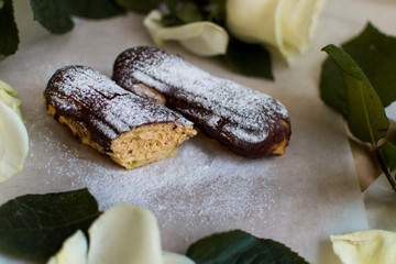 French eclairs with custard sprinkled with powdered sugar and white roses, petals, green leaves on light background