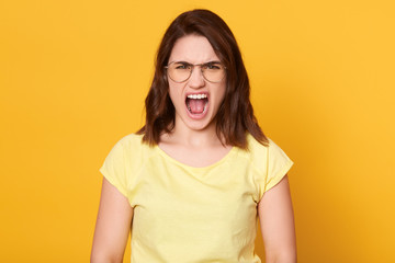 Horizontal shot of adorable woman with dark hair posing isolated over yellow background, attracyive lady looking directly at camera, screaming something, looks angry. People emotions concept.