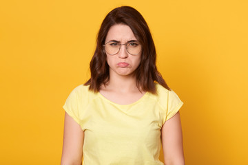 Obraz premium Close up portrait of beautiful young offended lady weraring casual t shirt and glasses, posing isolated over yellow background, woman expressers sadness, looks unhappy. People emotions cocnept.