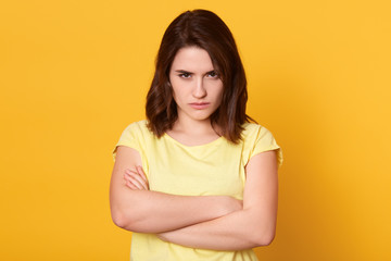 Obraz premium Close up portrait of beautiful dark haired woman wearing casual t shirt, standing and looks directly at camera with pouty lips, keeps hands folded, stands isolated on yellow background, being offended