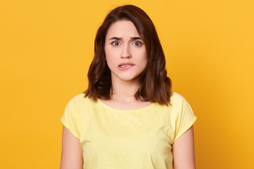 Obraz premium Close up portrait of nervous scared attractive brunette biting her lips, having frightened facial expression, having feeling of anxiety, wearing yellow t shirt, having short hair. Emotions concept.