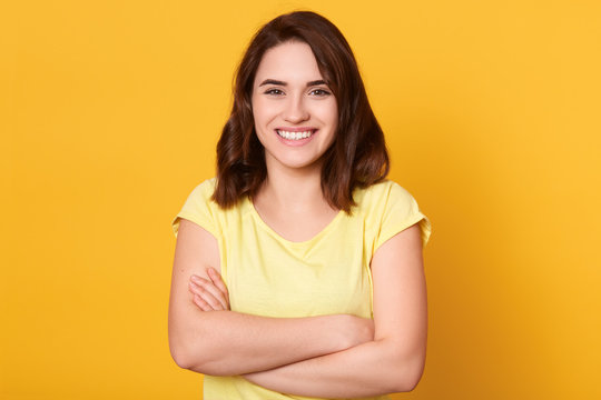 Studio shot of beautiful smiling woman with folded hands posing isolated over yellow background, looks happy, expresses joy, wearing casual clothing, having dark hair. People emotions concept.