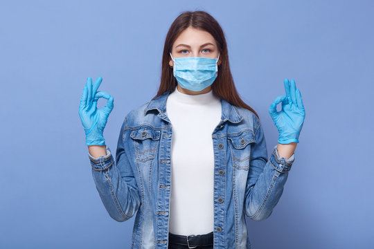 Picture of young woman showing sign ok, invoking to not panic, lady wearing protective face mask and disposable medical gloves, preventing virus infection, isolated over blue background. Coronavirus