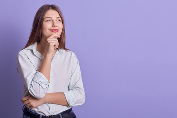 Fascinating blond business woman wearing white elegant blouse, looking aside with pencive facial expression, posing isoalted over lilac background. Copy space for adverisment or promotional text.