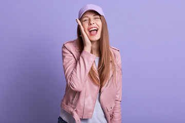 Naklejka premium Picture of attarctive woman dresses leather pink jacket and cap cavering ear with palm screaming something, expressing happyness, looks directly at camera, being in good mood. People emotions concept.