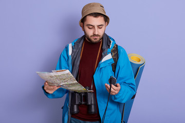 Indoor picture of experienced intrepid explorer holding map and compass in both hands, orientating in space, wearing cap, sweater and jacket, having binoculars around neck. Traveling concept.