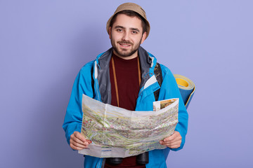 Portrait of handsome charismatic young tourist holding map in both hands, spending his free time in active way, wearing sweatshirt, jacket and hat, having sleeping pad aback. Orientation concept.