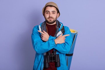 Studio shot of bearded guy wearing blue jacket and hat, holding rolled up karemat, points aside with both hands, posing isolated over purple studio background. Travelling and lifestyle concept.