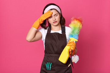 Close up portrait of tired housewife dresses brown apron, white casual t shirt and hair band, posing with cleaning deretgents isolated over pink background, having exuasted facial expressions.