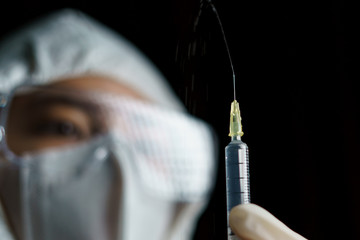 Woman wearing gloves with biohazard protective suit and mask holds an .virus vaccine with syringe,...