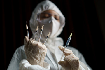 Woman wearing gloves with biohazard protective suit and mask holds an .virus vaccine with syringe,...