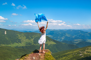 Beautiful young woman in white summer dress stands on a rock and looking into the amazing valley, blue scarf waving on the wind in her hands