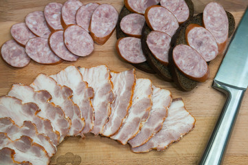 Rye bread sandwiches with smoked sausage and sliced bacon on wooden board. Rustic meat buffet top view.