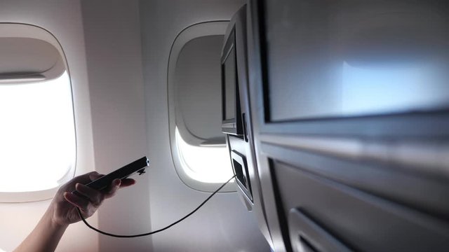 woman hand uses remote control for seatback TV set screen against bright porthole in airliner passenger cabin close view