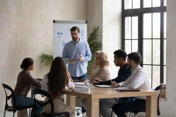 Confident male Caucasian team leader head meeting in office, present project on whiteboard, man speaker or coach talk make flip chart presentation to multiracial colleagues at briefing