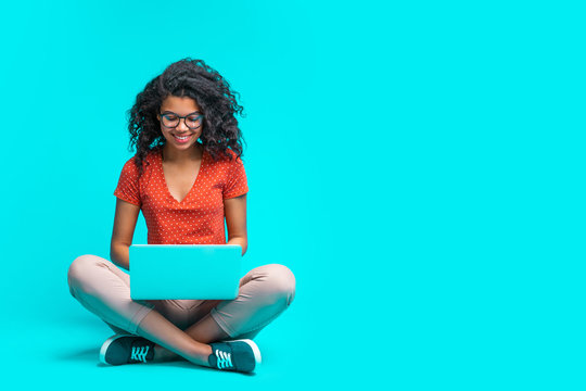Beautiful young smiling woman in casual outfit and trendy eyeglasses sitting isolated on bright colored blue background and working on her laptop