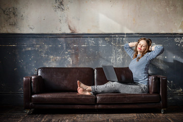 Obraz premium white young woman in jeans and a vest sitting on the sofa against the background of an old wall with a laptop and resting with a smile on her face, the sun rays