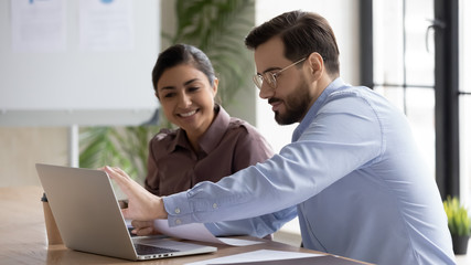 Smiling diverse businesspeople sit at desk work on laptop together discussing project or idea, happy multiracial colleagues cooperate using computer gadget, brainstorm at office meeting