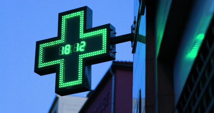 A pharmacy and illuminated international symbol hanging on a corner of a building with a twinkling clock in the middle of a green cross depicting a pharmacy caputred in 4k 60fps.