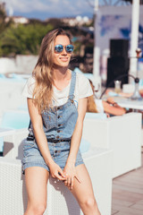 Satisfied traveler woman in casual clothes is sitting on the couch of an open cafe and posing enjoying the fresh sea air and the sun while letting go on a hot summer day