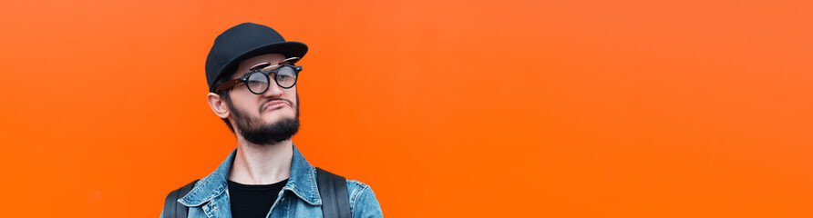 Obraz premium Panoramic banner, portrait of young confident hipster guy wearing denim jacket, round glasses and cap, isolated on background of lush lava color with copy space.
