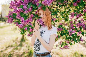 Handsome woman with red hair and pretty smile posing in lilac garden with flowers and enjoys the smell