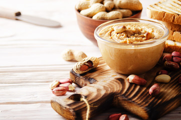 Low angle view at glass bowl with peanut butter on white wooden table with toasts aside. Healthy...