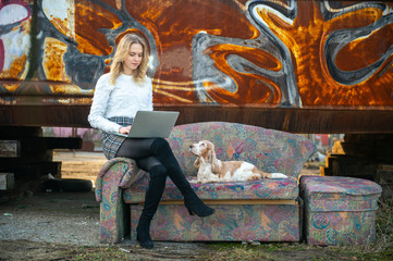 Cheerful caucasian young freelancer girl sitting on old couch outdoor over rusty graffiti wall on background and using on laptop with her lovely cocker spaniel dog lying close to her.