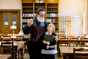 Attractive 70-aged grey-bearded grandfather in glasses showing the world of books in old vintage library for his smiling teen granddaughter, holding a book and listening her grandpa