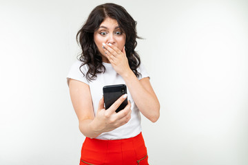 Fototapeta premium surprised girl covers her mouth with hand holding smartphone in front of herself on an isolated white background