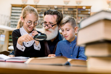 Two cheerful happy kids, boy and girl with magnifying glass glass listening to interesting book story from their handsome bearded grandfather or school teacher, sitting together in old library.
