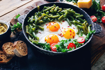 Pan of fresh fried eggs with tomatoes, green beans, bread, spices and herbs on rustic wooden background