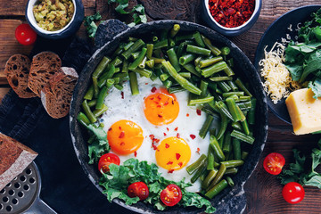 Pan of fresh fried eggs with tomatoes, green beans, bread, spices and herbs on rustic wooden background